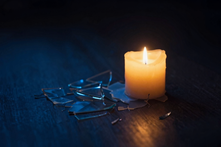 壊れたガラスの破片の隣に置かれた、少し溶けかけたろうそく。暗い部屋の中でかすかに灯っている。